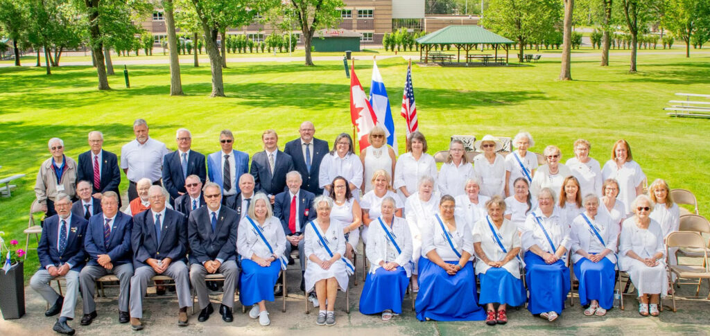 A group of people posing outdoors, seated and standing, dressed in formal and traditional attire with blue and white elements. Flags are displayed in the background, and a park with trees and a pavilion is visible.